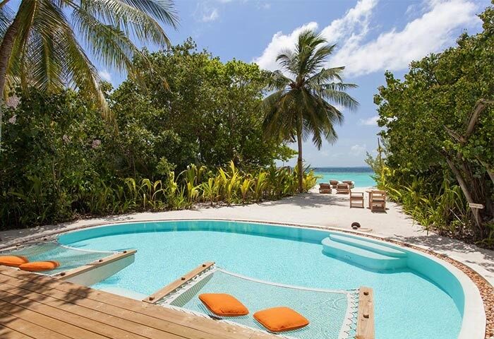 Soneva Fushi gay-friendly hotel pool view. Hammock with orange pillows in the pool. Tropical trees, beach, and ocean in the background.