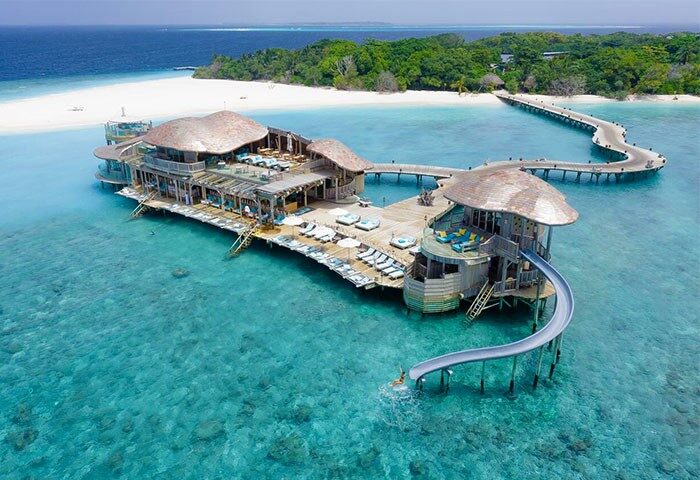 Gay-Friendly Hotel Soneva Fushi: Overwater villa with slide into turquoise water. Beach, island, and walkway in background. Luxury travel.