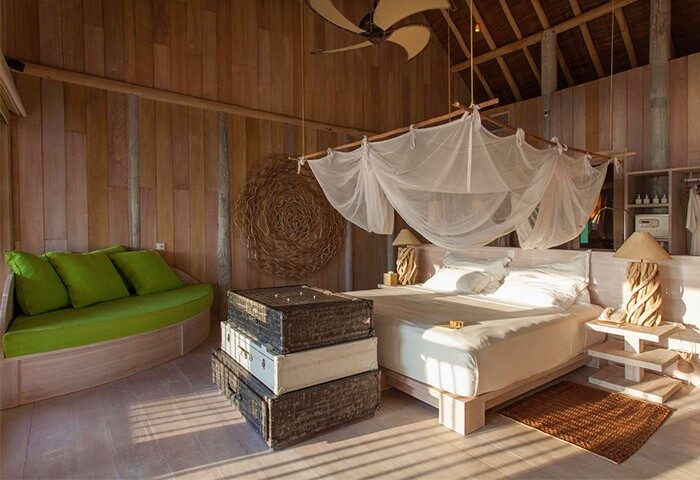 Soneva Fushi gay-friendly hotel room. Rustic wood interior with bed, mosquito net, green sofa, and stacked vintage suitcases. Luxury travel.