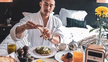 Gay man enjoys breakfast in bed at South Place Hotel, a pet-friendly hotel. He's wearing a robe and has toast, eggs, and juice.