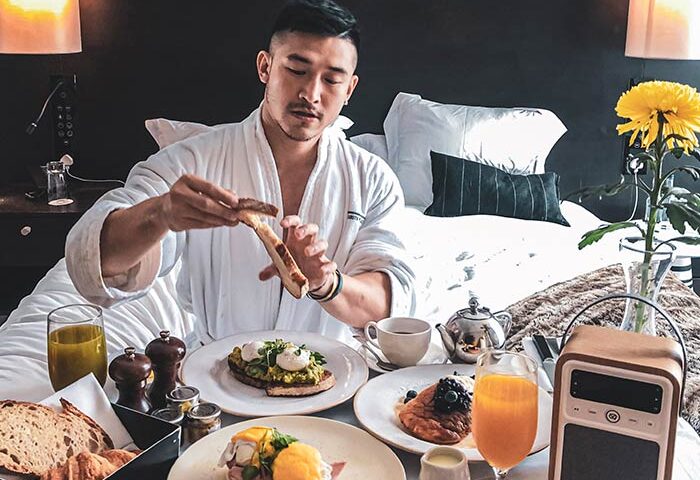 Gay man enjoys breakfast in bed at South Place Hotel, a pet-friendly hotel. He's wearing a robe and has toast, eggs, and juice.