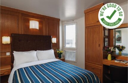 St Giles London Hotel room. Gay-friendly, pet-friendly hotel with double bed, blue striped duvet, wooden paneling, and We're Good To Go Visit England logo.