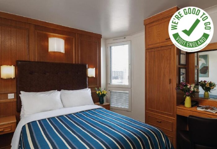 St Giles London Hotel room. Gay-friendly, pet-friendly hotel with double bed, blue striped duvet, wooden paneling, and We're Good To Go Visit England logo.