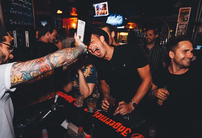 Gay-friendly St. Christopher's Hostel Berlin bar scene. Man with tattoos pouring drink into another man's mouth. Fun nightlife for gay travelers.