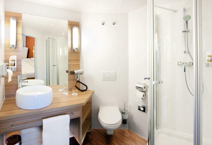 Star Inn Hotel Wien Schönbrunn bathroom. Modern design with shower, toilet, sink, and wood accents. Gay-friendly hotel in Vienna.