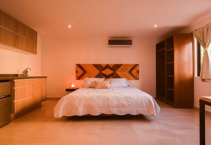 Stay Tropical gay-friendly hotel room. Queen bed with geometric headboard. Kitchenette, wardrobe, and window with curtains. Modern design.