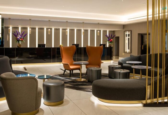 Lobby lounge at Strand Palace Hotel, a gay-friendly hotel. Modern seating area with chairs, sofas, and ottomans. Black reception desk with flowers.