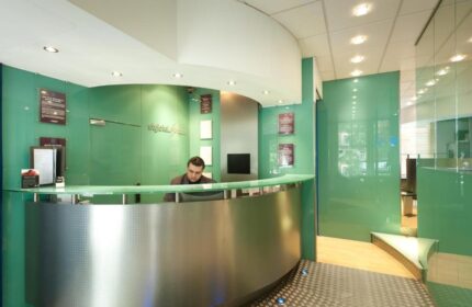 Stylotel, a gay-friendly hotel. Modern reception area with a man at the desk. Green glass walls and a curved, metallic front desk. Stylish, welcoming space.