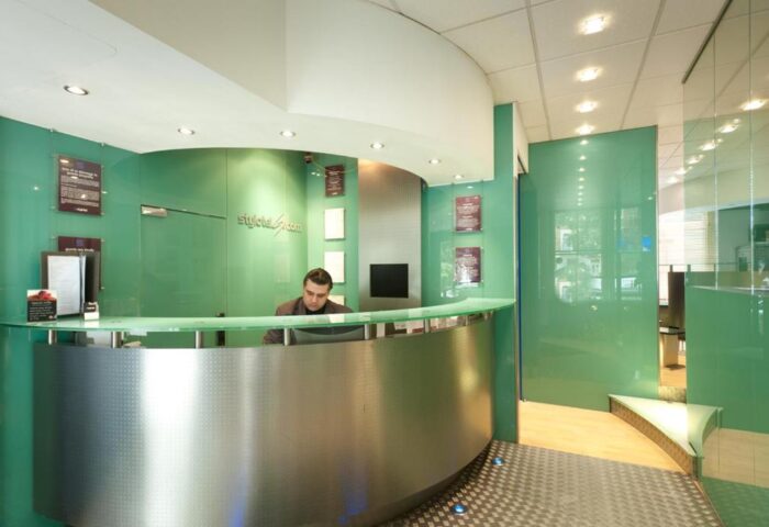 Stylotel, a gay-friendly hotel. Modern reception area with a man at the desk. Green glass walls and a curved, metallic front desk. Stylish, welcoming space.