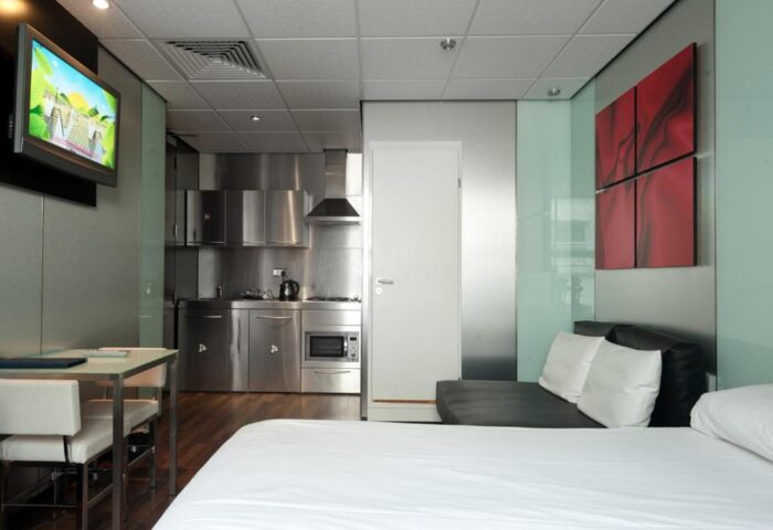 Stylotel, a gay-friendly hotel: Room interior with stainless steel kitchenette, bed, sofa, TV, table, chairs, and red artwork. Modern design.