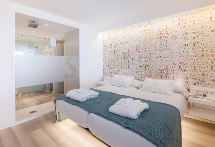 Sud Ibiza Suites gay-friendly hotel room. Double bed with robes, patterned wall, and glass-walled shower. Modern design for gay travelers.