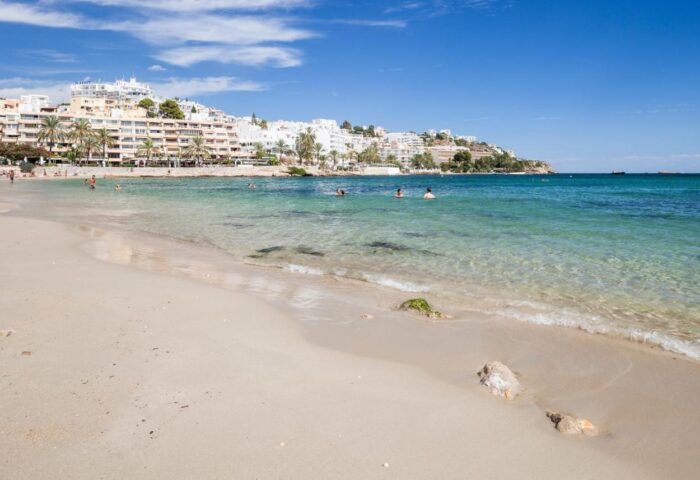 Beach view of Gay-Friendly Sud Ibiza Suites. Clear water, sandy beach, and hotel in the background. Ideal for gay men's travel advice.