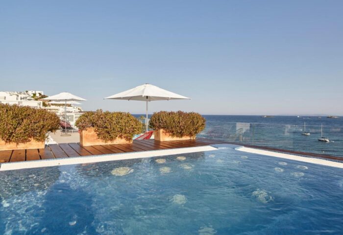 Sud Ibiza Suites gay-friendly hotel pool view. Ocean backdrop, umbrellas, and wood deck create a luxurious, inviting space.