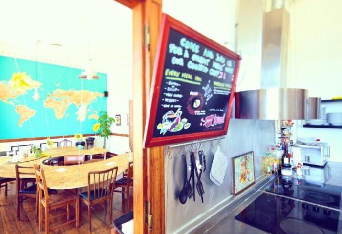 Sunset Destination Hostel's kitchen. Gay-friendly hotel with world map mural, communal dining table, and chalkboard menu. A welcoming space.