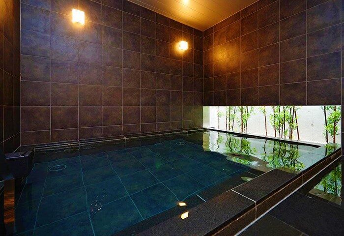 Kabukicho Super Hotel's bath. Dark tiled walls, lit by wall sconces, surround a deep soaking tub. Bamboo visible through a window. Gay travel.