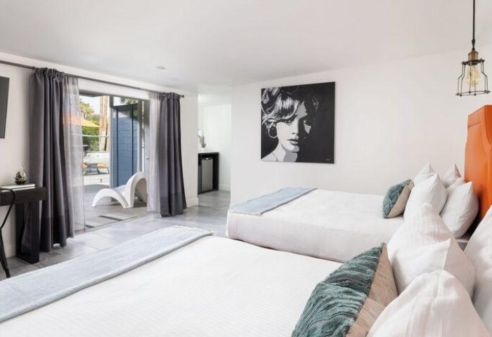 Gay-Friendly THE PALM SPRINGS HOTEL room. Two beds, white linens, blue accents, and modern art. Patio view with stylish outdoor seating.