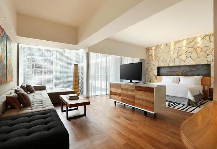 TS Suites Seminyak gay-friendly hotel room. Modern design with stone wall, wood floors, sofa, TV, and large windows. Bali luxury suite.