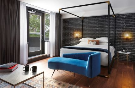 The Anndore House, part of JVD by Hyatt, gay-friendly hotel room. Features a four-poster bed, blue velvet couch, and exposed brick wall.