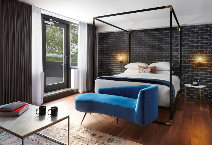 The Anndore House, part of JVD by Hyatt, gay-friendly hotel room. Features a four-poster bed, blue velvet couch, and exposed brick wall.