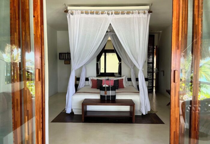 Bedroom view at The Beach Tulum Hotel, a gay-friendly hotel. Features a canopy bed, wooden accents, and a glimpse of the tropical surroundings.