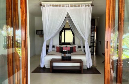 Bedroom view at The Beach Tulum Hotel, a gay-friendly hotel. Features a canopy bed, wooden accents, and a glimpse of the tropical surroundings.