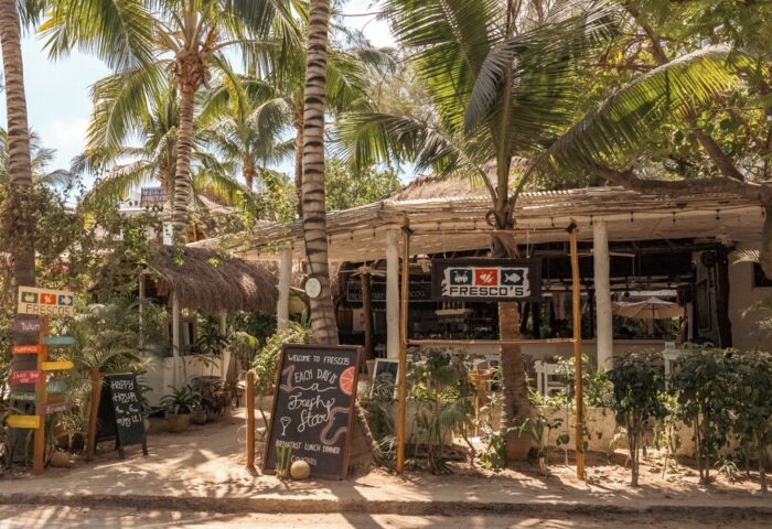 The Beach Tulum Hotel's gay-friendly Fresco's restaurant. Lush greenery, palm trees, and thatched roofs create a tropical vibe. Chalkboards advertise specials.