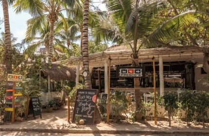 The Beach Tulum Hotel's gay-friendly Fresco's restaurant. Lush greenery, palm trees, and thatched roofs create a tropical vibe. Chalkboards advertise specials.