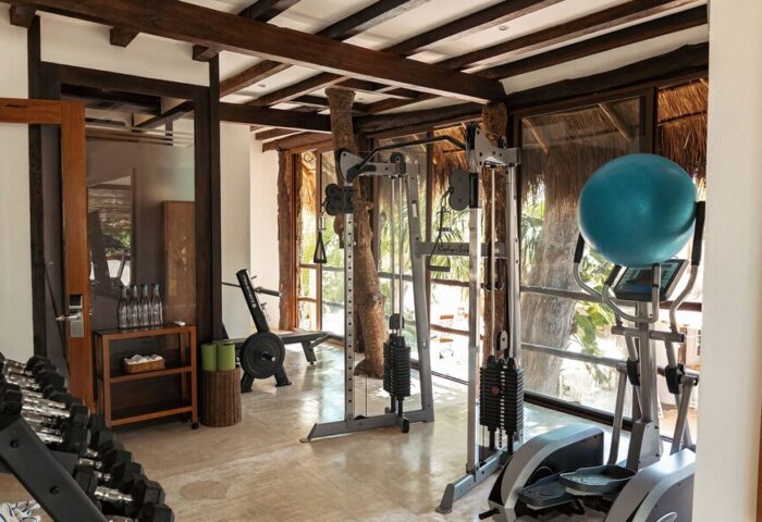 Gym at The Beach Tulum Hotel, a gay-friendly hotel. Exercise equipment, weights, and a blue exercise ball. Rustic wood beams and natural light.