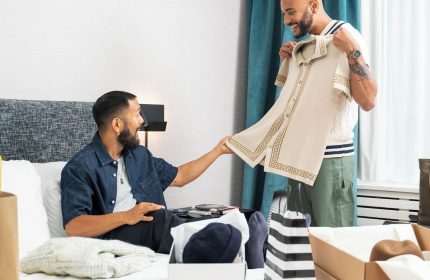 Two gay men at The Burrard Hotel, a gay-friendly hotel. One man shows the other a shirt. Shopping bags and clothing boxes are on the bed.