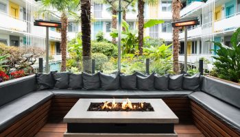 The Burrard Hotel's gay-friendly courtyard. Outdoor seating area with a fire pit, lush greenery, and colorful hotel balconies. A relaxing space.