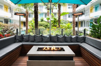 The Burrard Hotel's gay-friendly courtyard. Outdoor seating area with a fire pit, lush greenery, and colorful hotel balconies. A relaxing space.