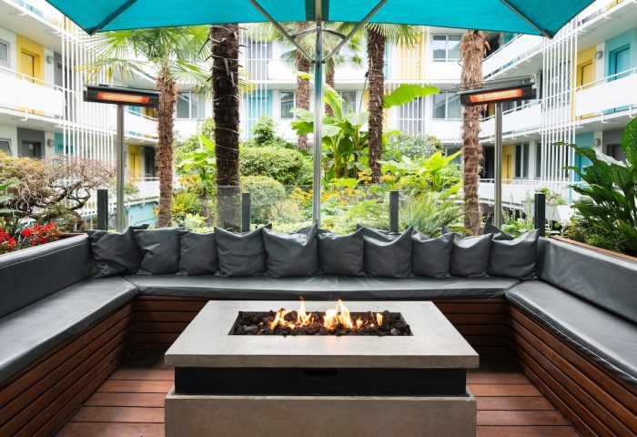 The Burrard Hotel's gay-friendly courtyard. Outdoor seating area with a fire pit, lush greenery, and colorful hotel balconies. A relaxing space.