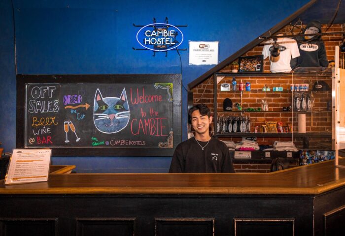 Cambie Hostel Gastown's reception desk with a smiling staff member. A chalkboard displays bar specials. Gay-friendly hotel for your travel advice.