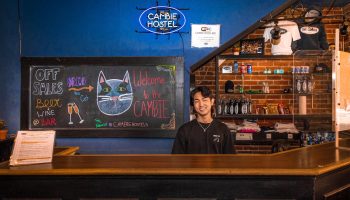 Cambie Hostel Gastown's reception desk with a smiling staff member. A chalkboard displays bar specials. Gay-friendly hotel for your travel advice.