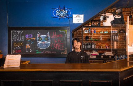 Cambie Hostel Gastown's reception desk with a smiling staff member. A chalkboard displays bar specials. Gay-friendly hotel for your travel advice.
