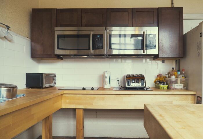 Kitchen at The Cambie Hostel Gastown, a gay-friendly hotel. Microwaves, toaster, and kettle on a wooden countertop. Brown cabinets above.