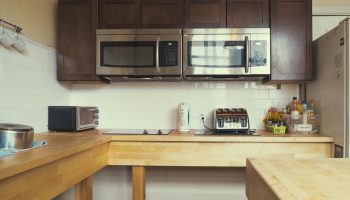 Kitchen at The Cambie Hostel Gastown, a gay-friendly hotel. Microwaves, toaster, and kettle on a wooden countertop. Brown cabinets above.