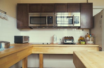 Kitchen at The Cambie Hostel Gastown, a gay-friendly hotel. Microwaves, toaster, and kettle on a wooden countertop. Brown cabinets above.