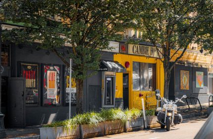 Cambie Hostel Gastown's exterior. This gay-friendly hotel features a motorcycle parked outside. A vibrant yellow facade stands out.