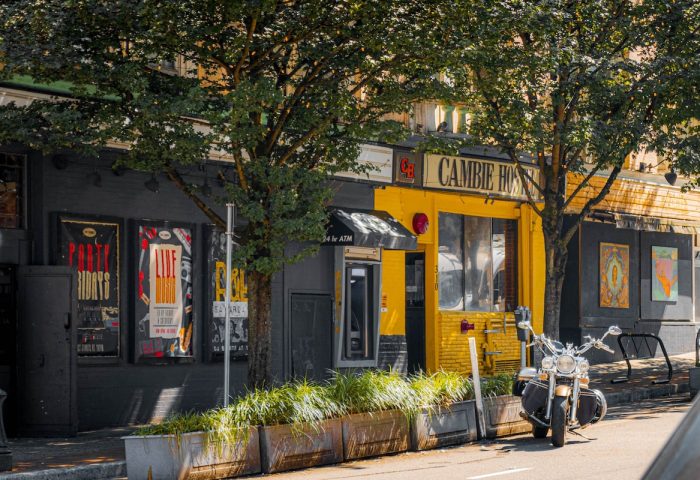 Cambie Hostel Gastown's exterior. This gay-friendly hotel features a motorcycle parked outside. A vibrant yellow facade stands out.