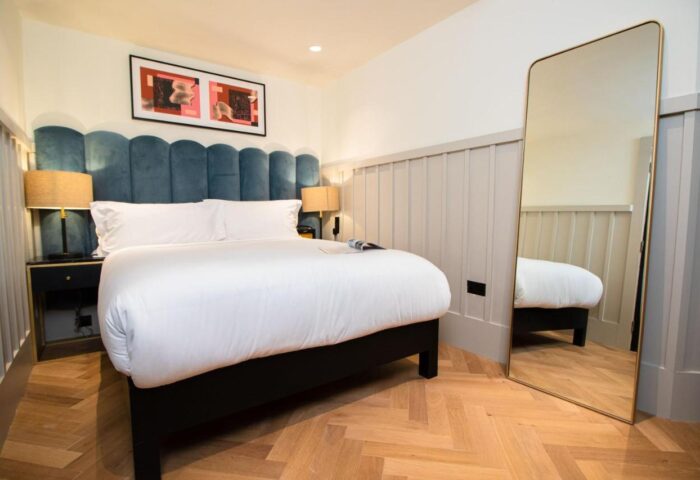 The Clarence gay-friendly hotel room. Bed with blue headboard, white linens, bedside tables, lamps, art, full-length mirror, and wood floor.