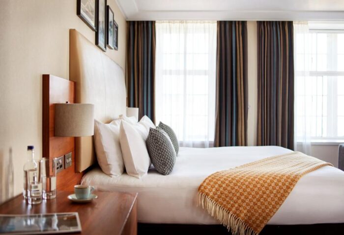 The Clermont London Charing Cross gay-friendly hotel bedroom. Double bed, pillows, bedside table with lamp, water, and coffee. Curtains on windows.