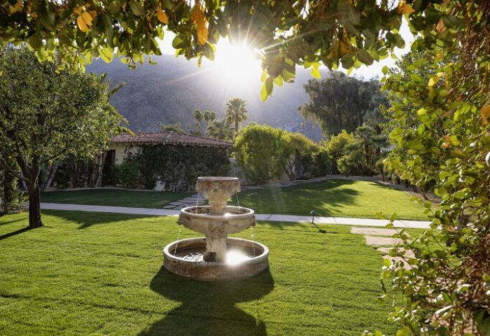 Colony Palms Hotel fountain in a lush garden. Gay-friendly hotel with sunshine, green grass, and mountain views. Palm Springs gay travel.