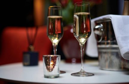 Champagne flutes at The Dilly, a gay-friendly hotel. Sparkling wine, candle, and ice bucket on a white table suggest luxury travel.