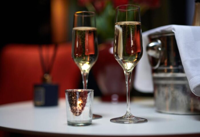 Champagne flutes at The Dilly, a gay-friendly hotel. Sparkling wine, candle, and ice bucket on a white table suggest luxury travel.