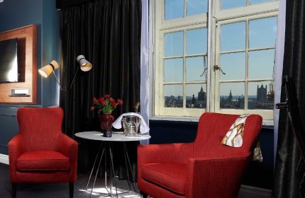 The Dilly Hotel's gay-friendly room features red chairs, a table with champagne, and a London cityscape view. Ideal gay travel.