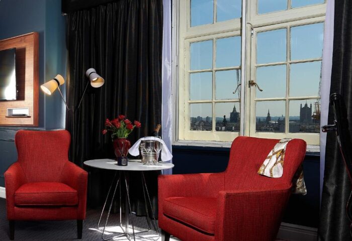 The Dilly Hotel's gay-friendly room features red chairs, a table with champagne, and a London cityscape view. Ideal gay travel.