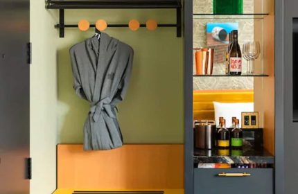 Interior of the Drake Hotel, a gay-friendly hotel. A robe hangs over a bench. A shelf displays a bottle of wine and glasses.