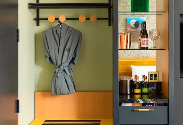 Interior of the Drake Hotel, a gay-friendly hotel. A robe hangs over a bench. A shelf displays a bottle of wine and glasses.