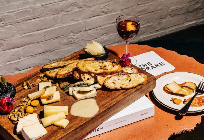The Drake Hotel's gay-friendly ambiance: A charcuterie board with cheese, bread, nuts, and a cocktail. Cozy fireplace setting, perfect for gay travelers.